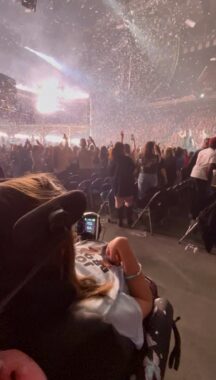 A young woman sits in her wheelchair on the floor of an arena during a Jonas Brothers concert. We can't quite see the stage, but there are bright lights shining and confetti raining down on the audience.