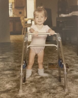 A classic '80s photograph shows a toddler in a leotard smiling as she holds onto a small walker in the living room of an '80s-style home complete with brown carpet. 