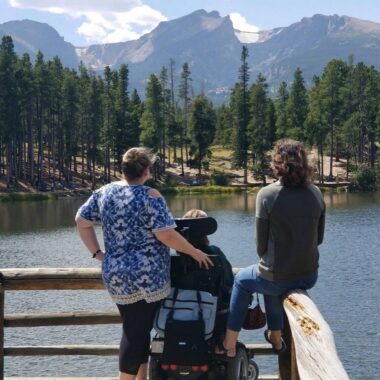 Three people on a dock look out over a lake to mountains in the distance.